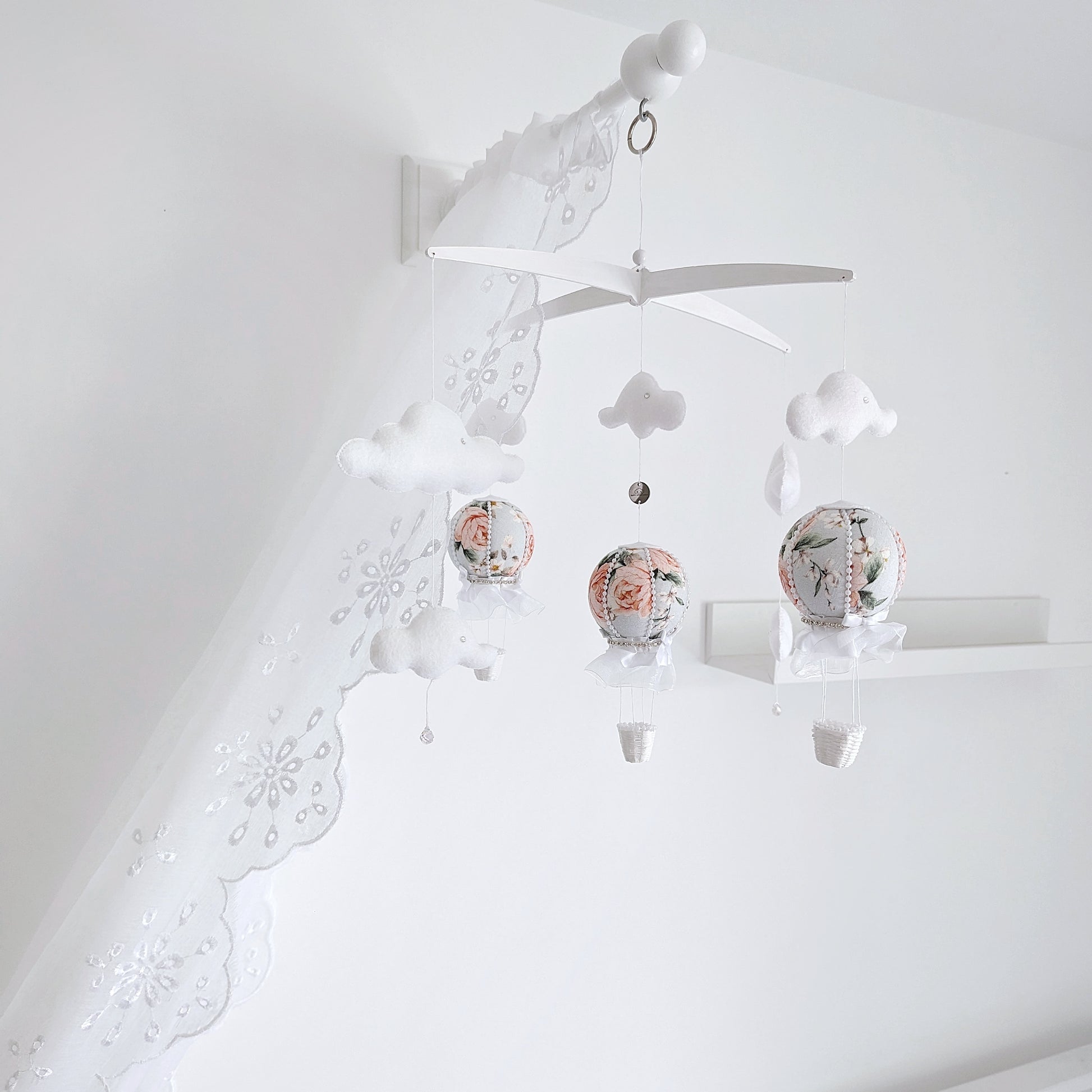 Delicate handmade baby mobile with floral hot air balloons, soft clouds, and a cross hanger in a white nursery.